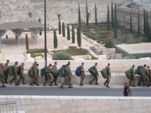 IDF soldiers march along a road passing a civilian.