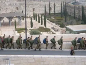 IDF soldiers march along a road passing a civilian.