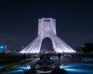 An image taken at night of the Azadi Tower also known as the Freedom Tower in center of Ian's capital, Tehran. The building is lit up with white lights against a black sky.