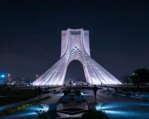An image taken at night of the Azadi Tower also known as the Freedom Tower in center of Ian's capital, Tehran. The building is lit up with white lights against a black sky.