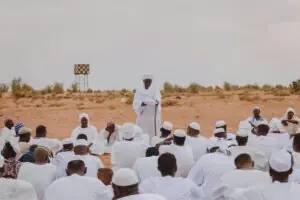 Gathering of men dressed in white listening to a male speaker in the Sudanese desert.
