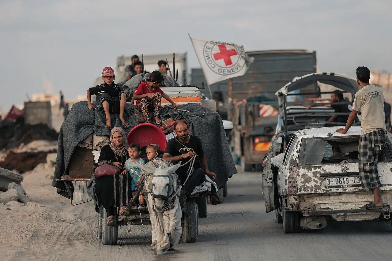 Displaced Palestinians moving in a convey via horse and cart. A Red Cross flag flies in the background.