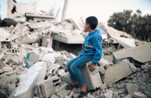 A child in Gaza dressed in a blue tracksuit suits on top of a mound of rubble and staring at the destruction.