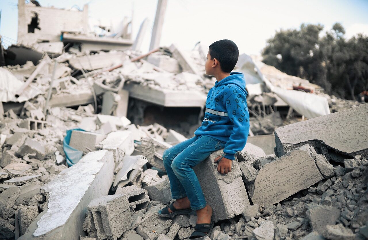 A child in Gaza dressed in a blue tracksuit suits on top of a mound of rubble and staring at the destruction.