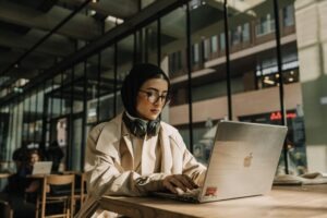 Women in a cream coat and a black headscarf sits in a library typing on her laptop